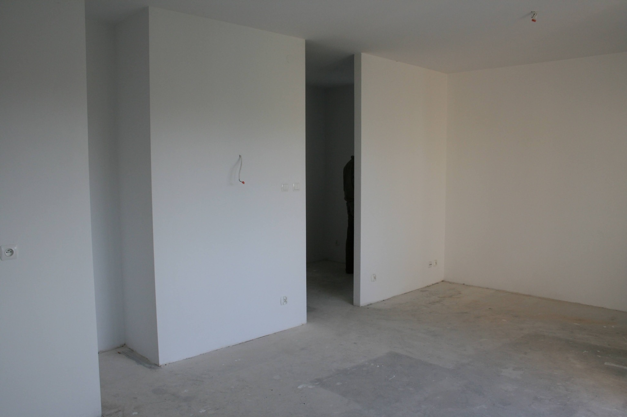 Surowe wnętrze mieszkania w stanie deweloperskim. Białe ściany, betonowa podłoga, widoczne instalacje elektryczne. Minimalistyczny kadr z perspektywą na kolejne pomieszczenia.