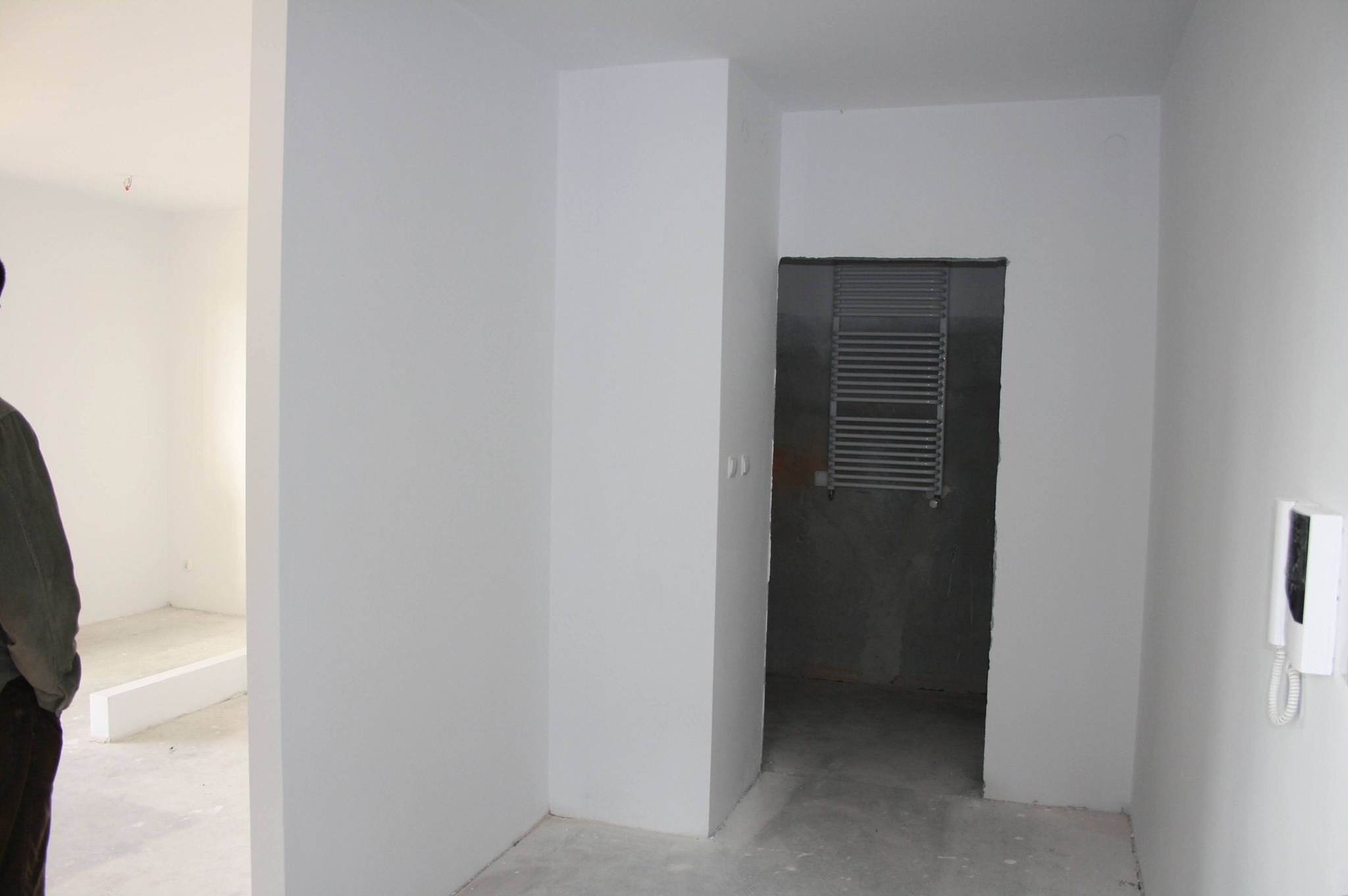 Wnętrze w stanie deweloperskim: białe ściany, surowa podłoga, grzejnik drabinkowy w łazience. Widoczny domofon i fragment osoby po lewej stronie kadru.