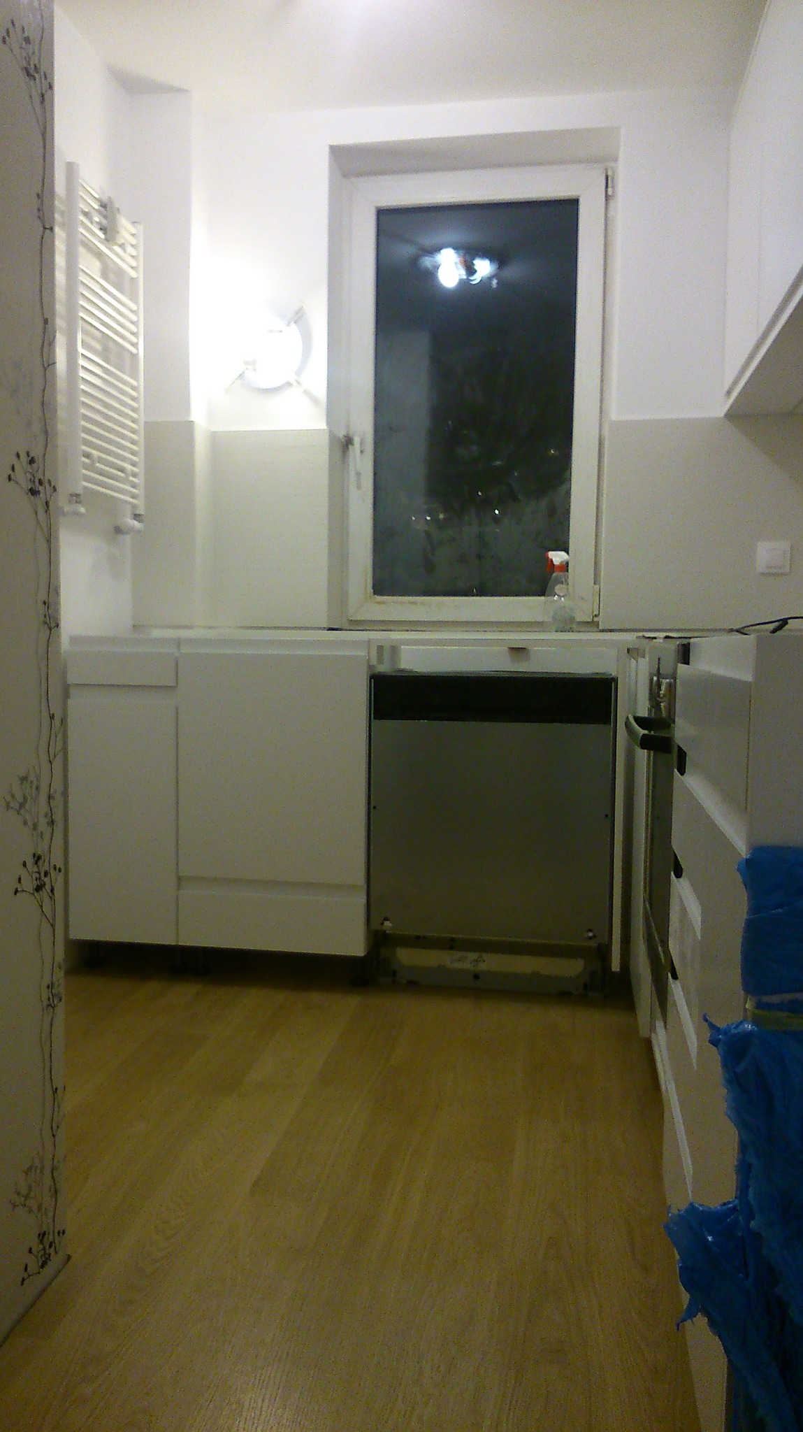 Kuchnia w trakcie montażu: białe szafki, drewniana podłoga, okno z widokiem na nocne miasto. Widoczna niedokończona zabudowa zmywarki i elementy instalacji.