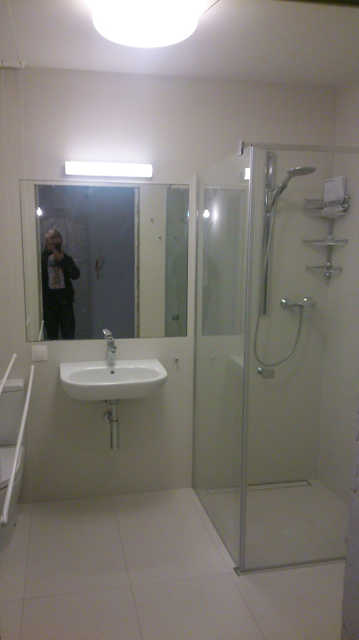 Jasna łazienka z kabiną prysznicową i umywalką. W lustrze odbija się osoba robiąca zdjęcie. Minimalistyczny design, białe płytki podłogowe, proste oświetlenie.