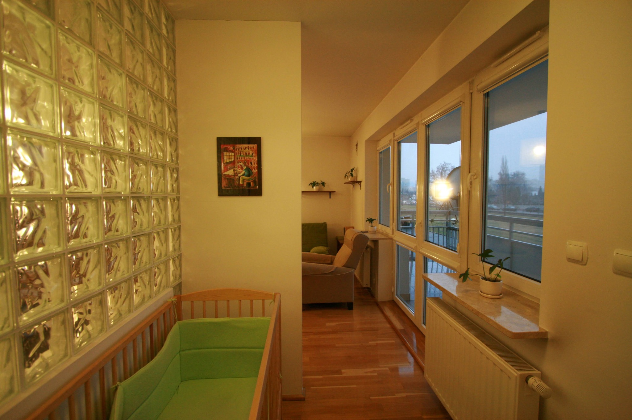 Umeblowane mieszkanie 36m2 po remoncie: pokój dziecięcy z łóżeczkiem, ściana z luksferów, widok na salon i balkon z roślinami w doniczkach na parapecie.