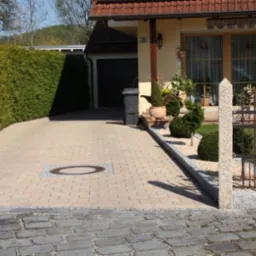 Jasna kostka brukowa na podjeździe do domu z widoczną studzienką, obok zadbany żywopłot i ozdobne krzewy przy ogrodzeniu.