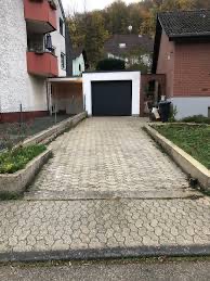 Podjazd do garażu wyłożony szarą kostką brukową o wzorze plastra miodu, obrzeża z betonowych krawężników, w tle domy jednorodzinne i drzewa.