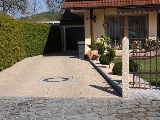 Jasna kostka brukowa na podjeździe do domu z widoczną studzienką, obok zadbany żywopłot i ozdobne krzewy przy ogrodzeniu.