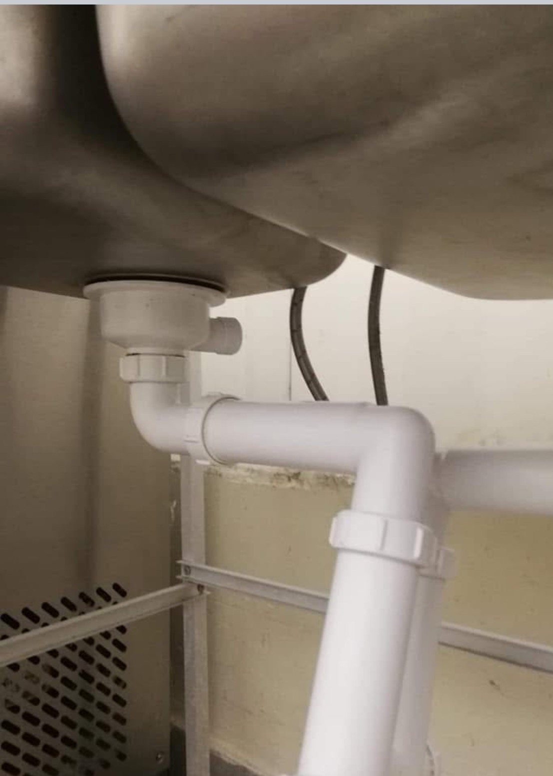 Widok od dołu na odpływ zlewozmywaka ze stali nierdzewnej z białymi rurami PCV i metalową konstrukcją wsporczą w tle.