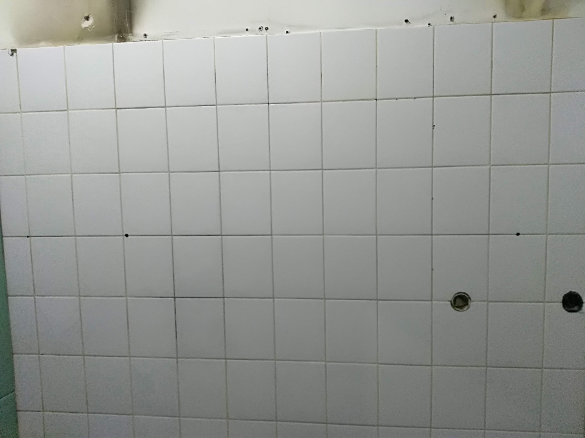 Ściana z białych, kwadratowych płytek ceramicznych z nawierconymi otworami, widoczne ślady po demontażu elementów.