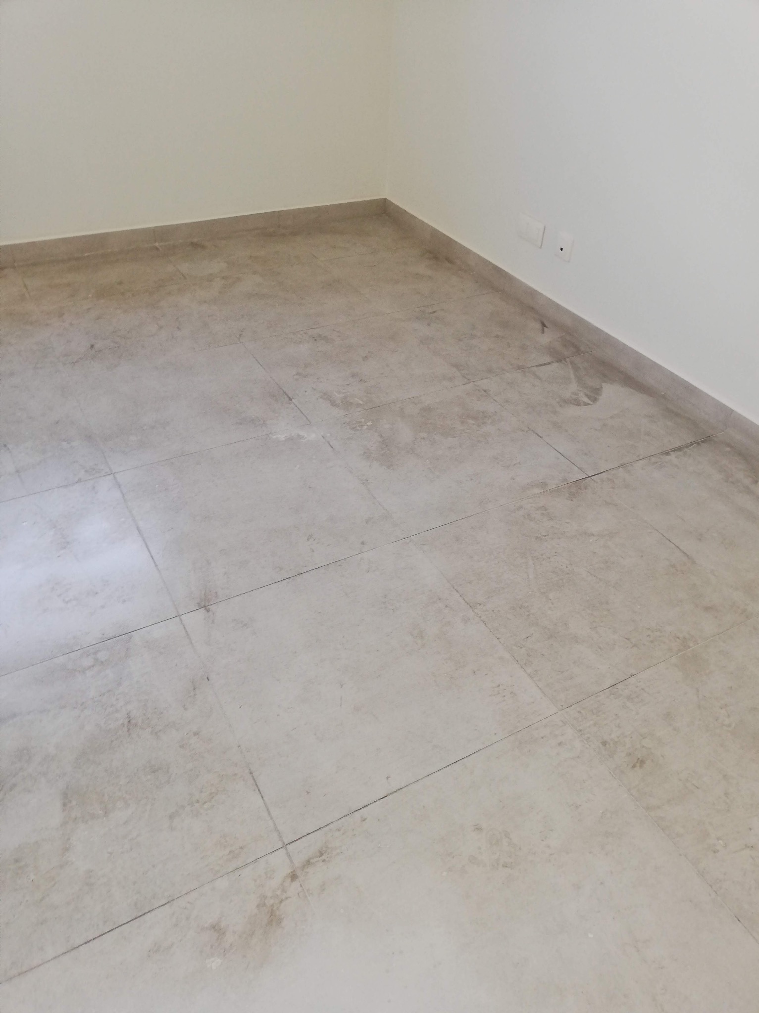 Pusta przestrzeń z beżowymi płytkami podłogowymi, widoczne zabrudzenia na fugach i powierzchni płytek, białe ściany z gniazdkami elektrycznymi.