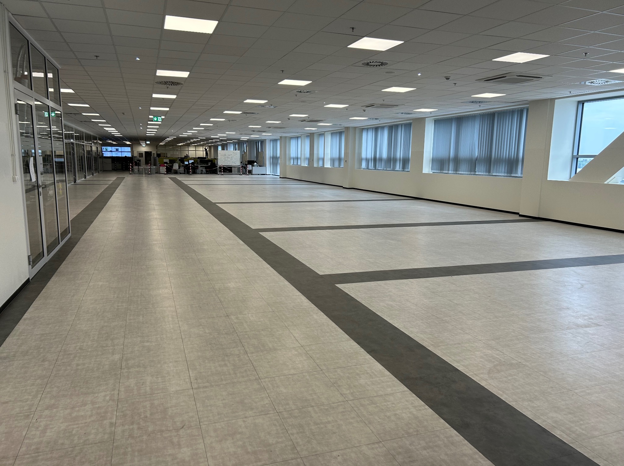 Przestronne, puste biuro z jasną podłogą w płytkach i ciemnoszarymi pasami, rzędy okien z żaluzjami i sufit z kwadratowymi lampami, widok z perspektywy podłogi.