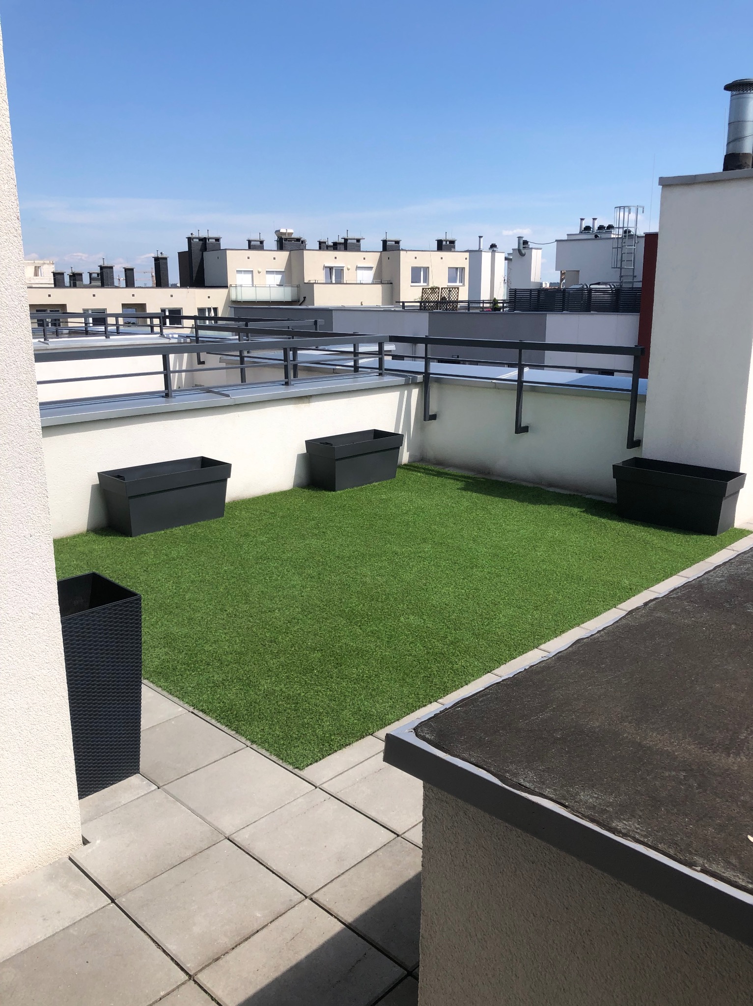 Widok na taras na dachu z sztuczną trawą, czarnymi donicami i szarymi płytami chodnikowymi w słoneczny dzień.
