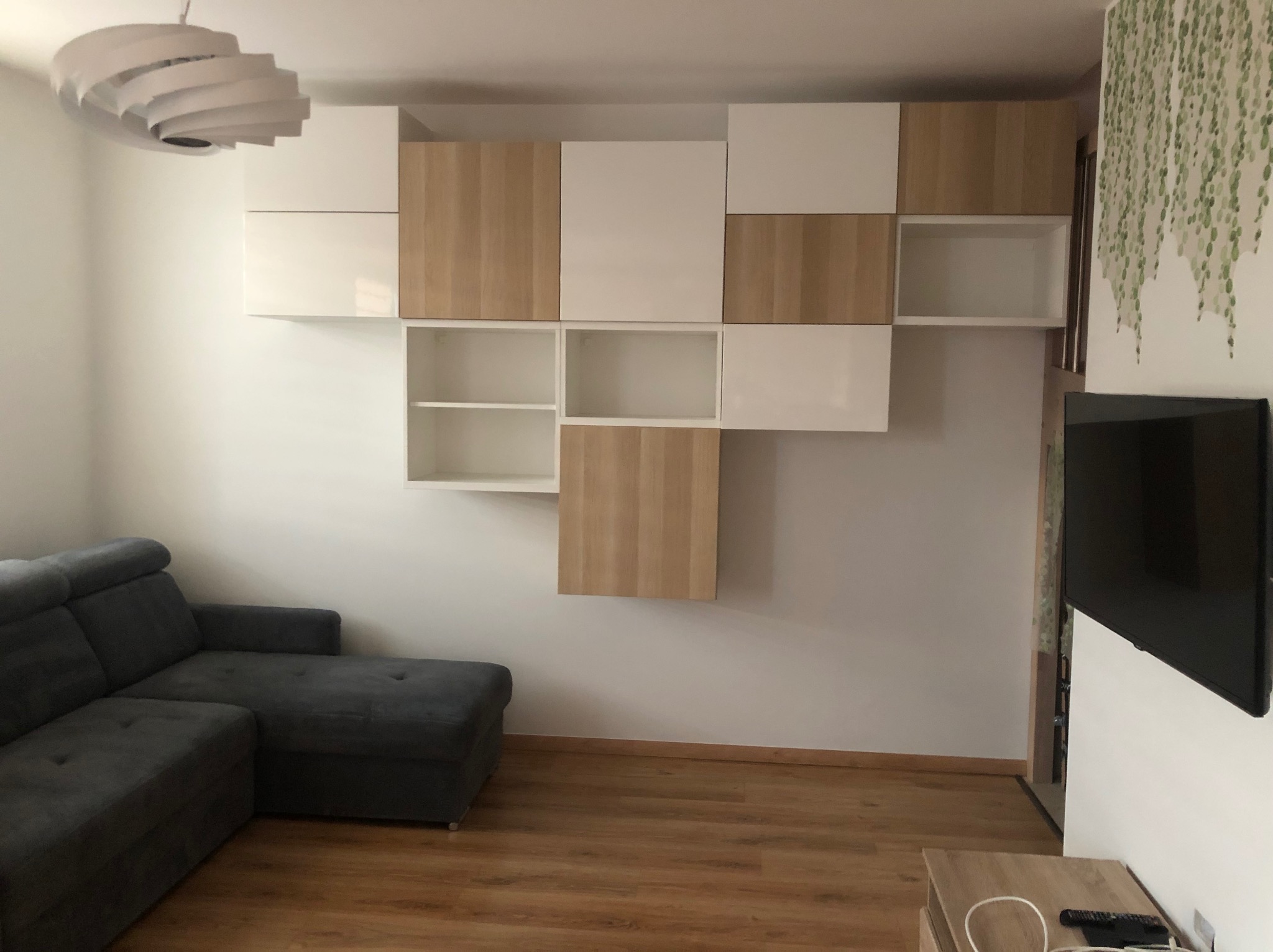 Jasne wnętrze salonu z szarą sofą narożną, modułowymi szafkami wiszącymi w kolorze białym i drewnopodobnym oraz dekoracją ścienną z motywem roślinnym.