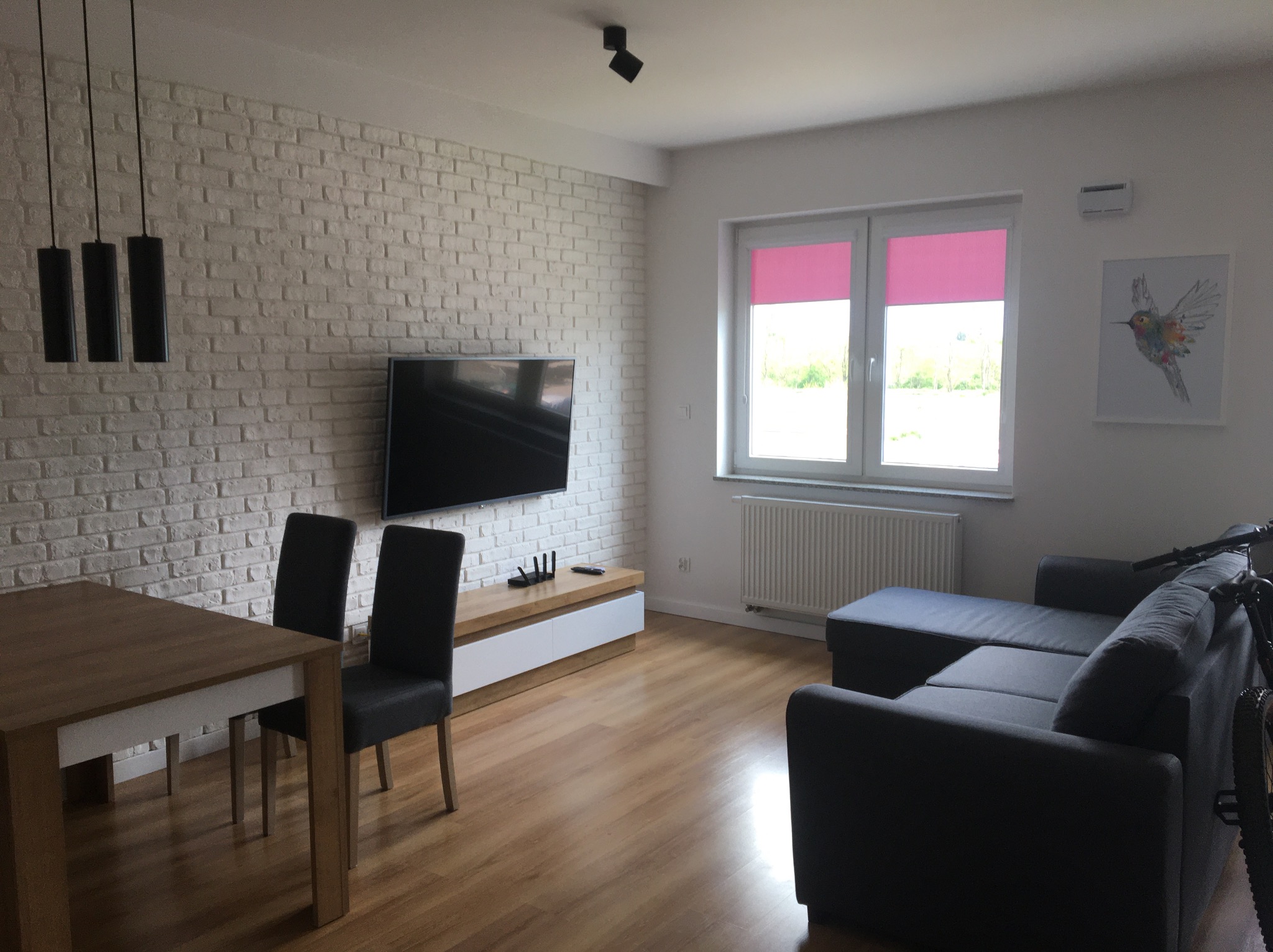 Jasne wnętrze salonu z ceglaną ścianą, drewnianym stołem i szarą sofą narożną; różowe rolety w oknie.