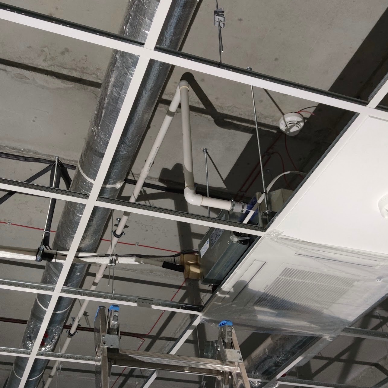 Widok instalacji pod sufitem podwieszanym: rury PCV, kanały wentylacyjne w izolacji, czujnik dymu i klimatyzator kasetonowy w folii ochronnej, z widoczną drabinką monterską.