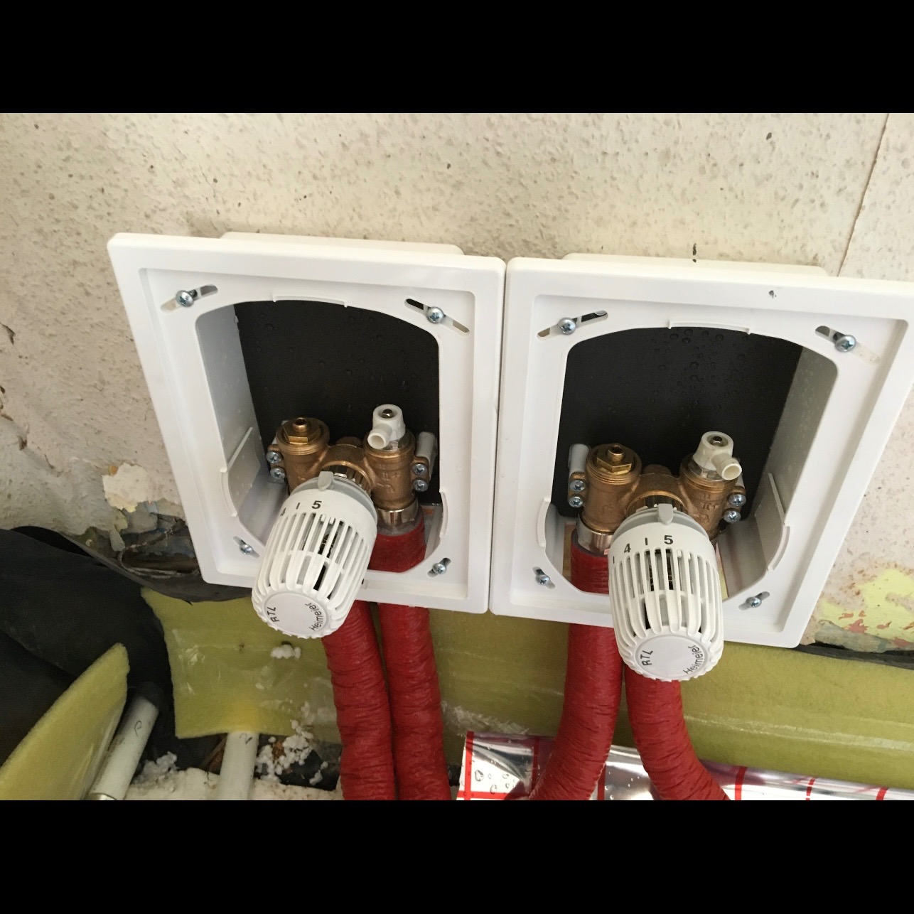 Dwa zawory termostatyczne w białych obudowach, zamontowane przy czerwonych rurach instalacji grzewczej, widoczne ślady prac montażowych na ścianie.