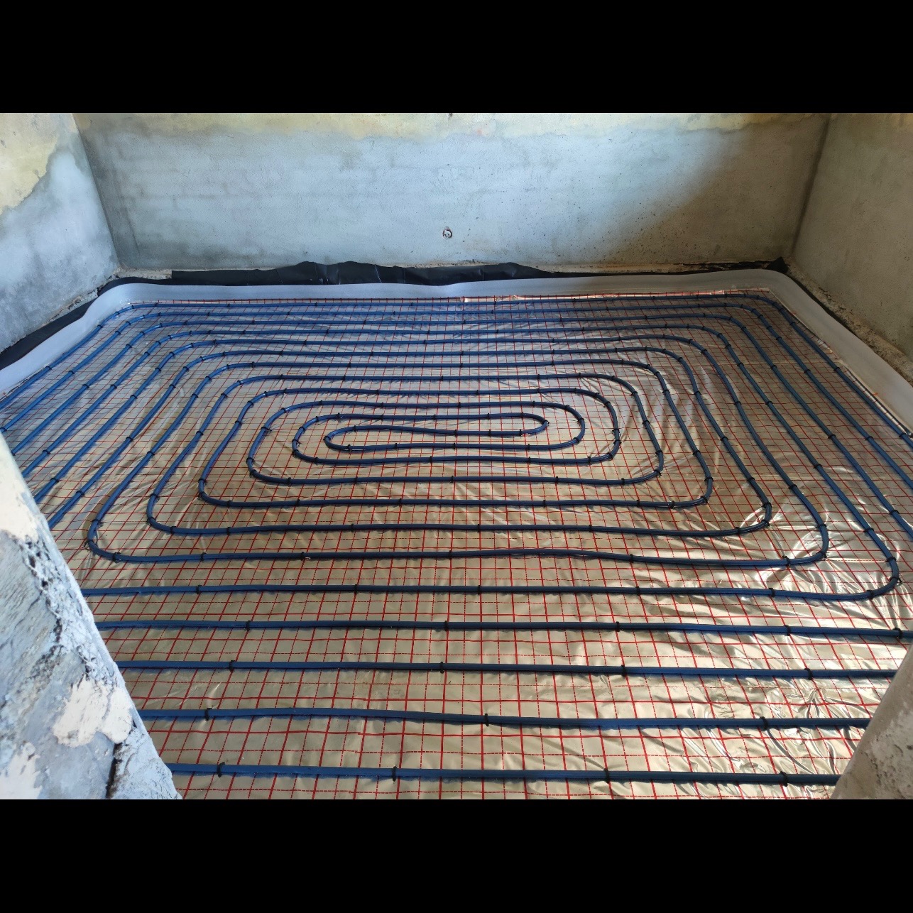 Niebieskie rury ogrzewania podłogowego ułożone spiralnie na folii z czerwoną siatką w surowym pomieszczeniu z betonowymi ścianami.