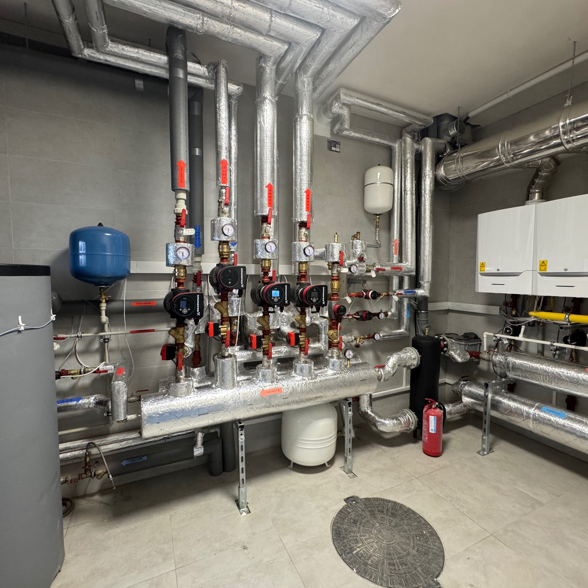 Węzeł cieplny z pompami obiegowymi, manometrami i zabezpieczeniami, rury w izolacji aluminiowej, zbiorniki wyrównawcze, kocioł gazowy, gaśnica w pomieszczeniu technicznym z szarą płytką na ścianach...