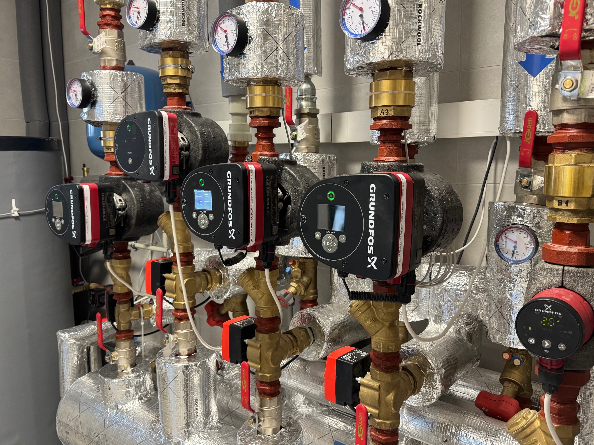 Złożona instalacja grzewcza z pompami obiegowymi Grundfos, zaworami, manometrami i izolacją termiczną, widoczne liczne rury miedziane i stalowe.