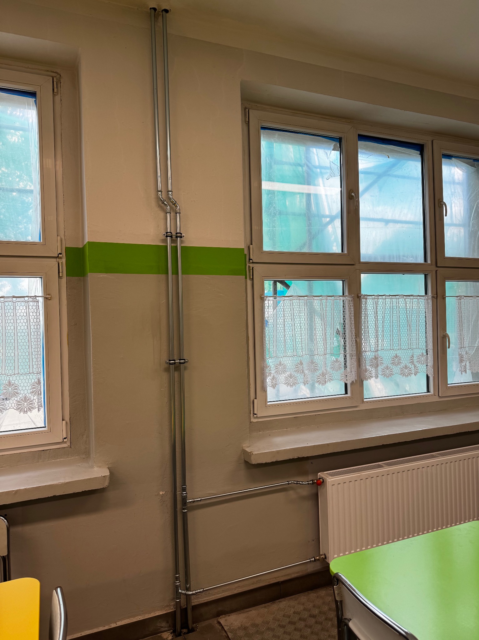 Dwie pionowe rury instalacyjne w metalowej osłonie, przymocowane do ściany w pomieszczeniu z oknami, grzejnikiem i fragmentem stołu z krzesłem. Widoczny fragment instalacji grzewczej z grzejnikiem.