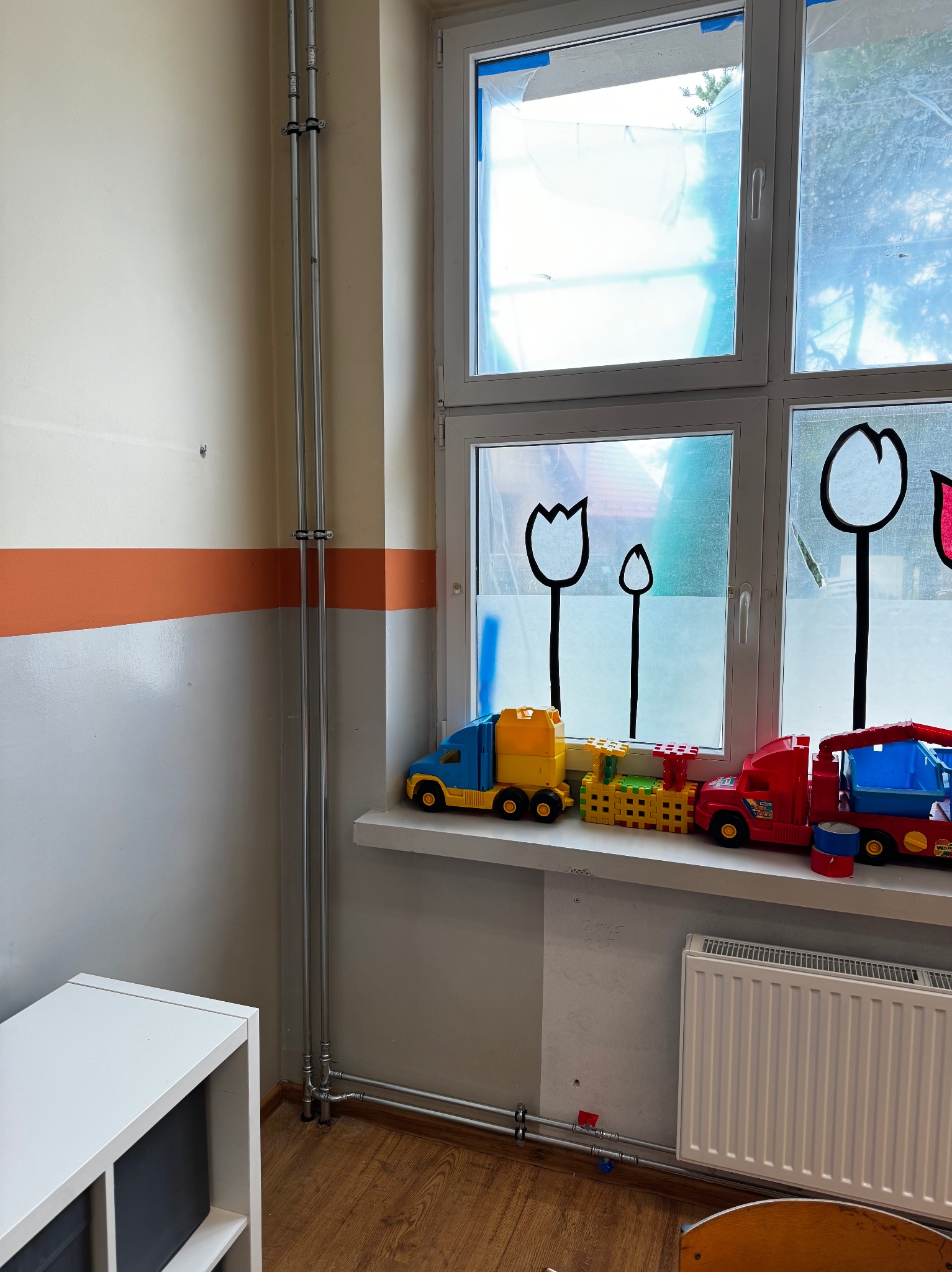Dwie chromowane rury pionowe instalacji grzewczej, biegnące wzdłuż ściany z pomalowanym na pomarańczowo pasem, obok okna z naklejonymi konturami tulipanów i zabawkami na parapecie, nad grzejnikiem.