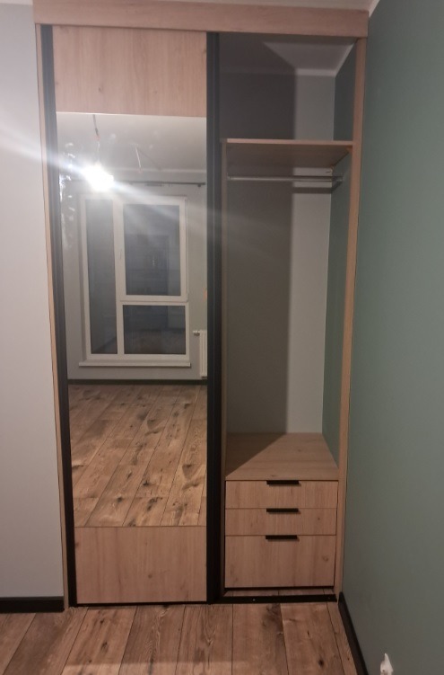 Wnętrze szafy wnękowej z przesuwnymi drzwiami lustrzanymi, widoczne półki, drążek na ubrania i trzy szuflady w kolorze jasnego drewna.