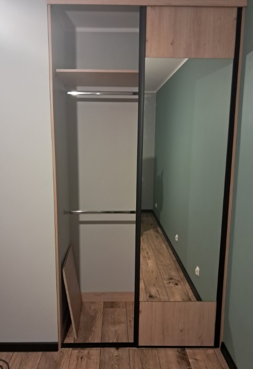 Wnętrze szafy przesuwnej z lustrem, widoczne półki, drążki na ubrania i fragment podłogi z paneli w odbiciu lustra.