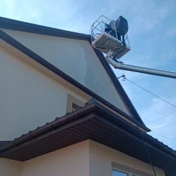 RClean - Pracownicy na podnośniku koszowym, w trakcie czyszczenia jasnej elewacji domu jednorodzinnego z brązowym dachem, widoczne smugi po myciu.