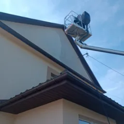 Pracownicy na podnośniku koszowym, w trakcie czyszczenia jasnej elewacji domu jednorodzinnego z brązowym dachem, widoczne smugi po myciu.