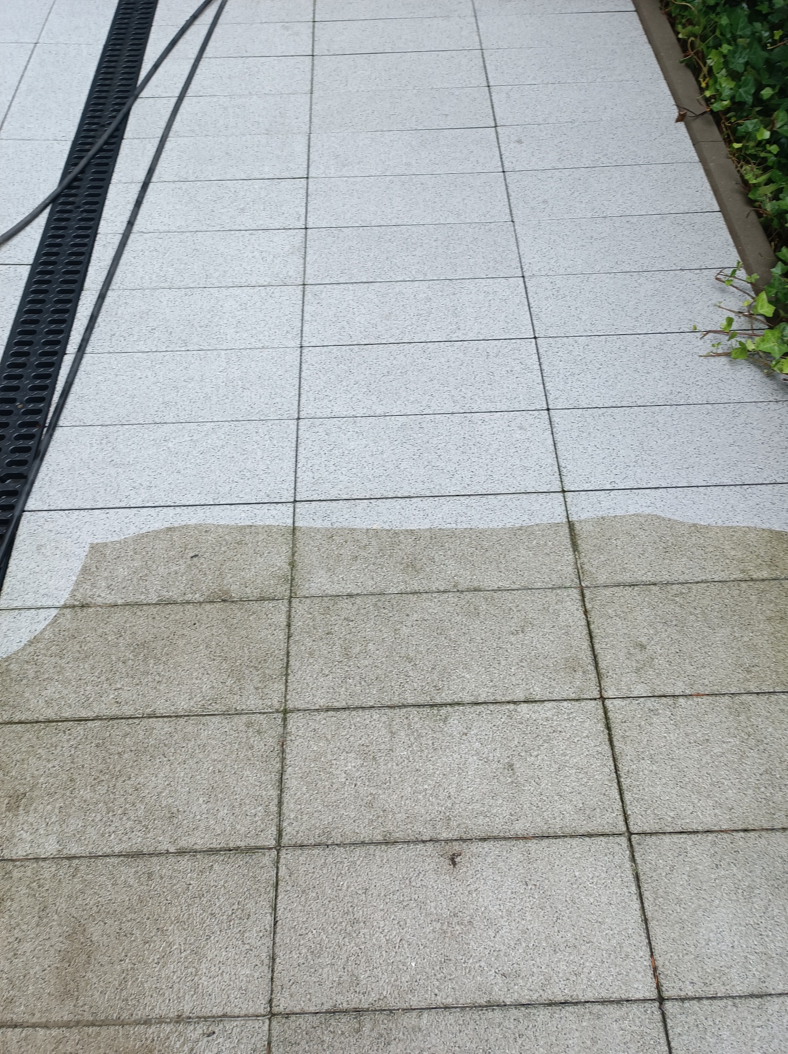 Częściowo umyta szara kostka brukowa z widoczną różnicą w kolorze, czarna kratka odpływowa po lewej stronie, fragment zieleni przy krawędzi po prawej.