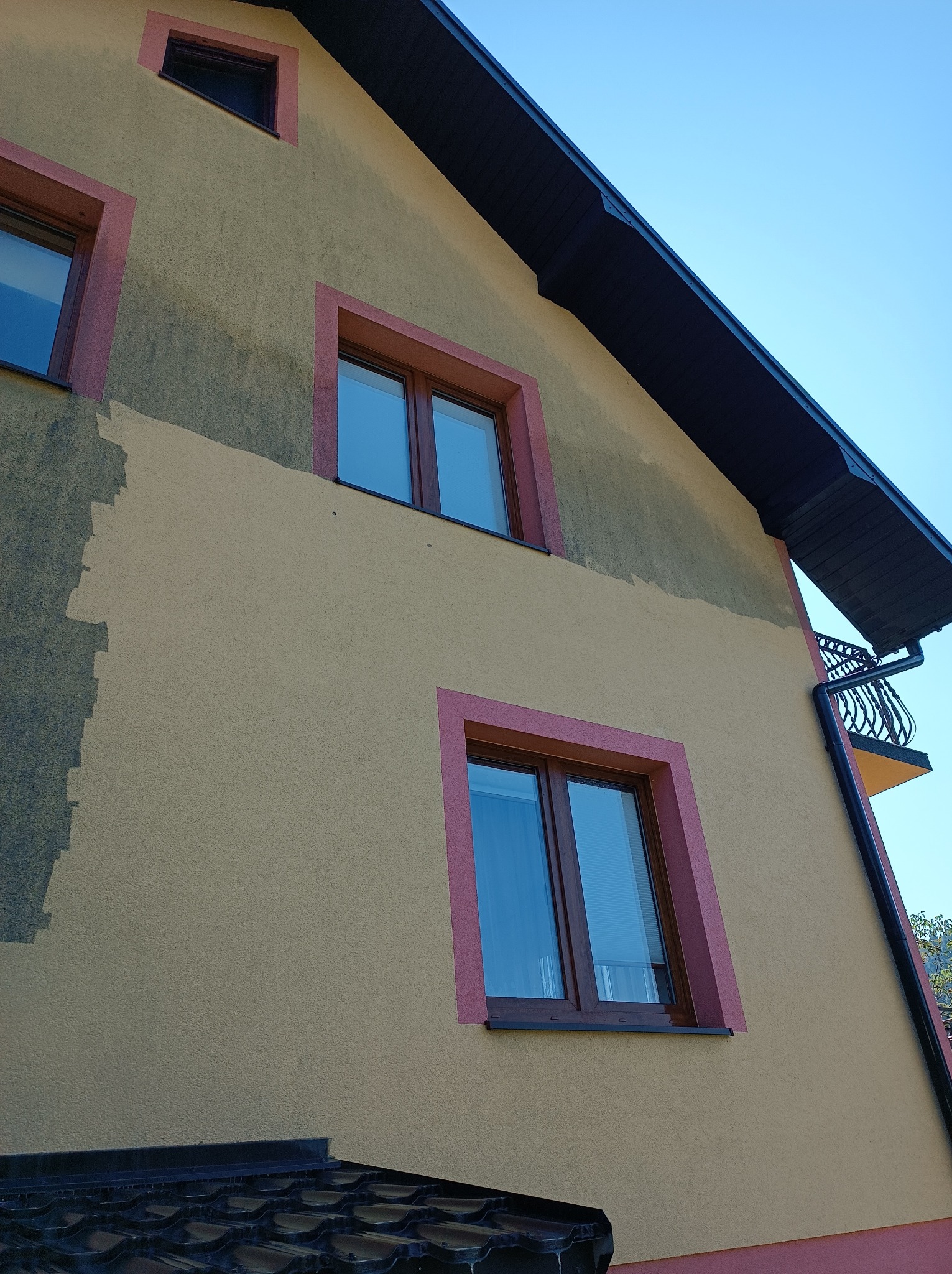 Fragment elewacji budynku z widocznym obszarem po czyszczeniu, kontrastującym z zabrudzoną powierzchnią, okna w bordowych ramach.
