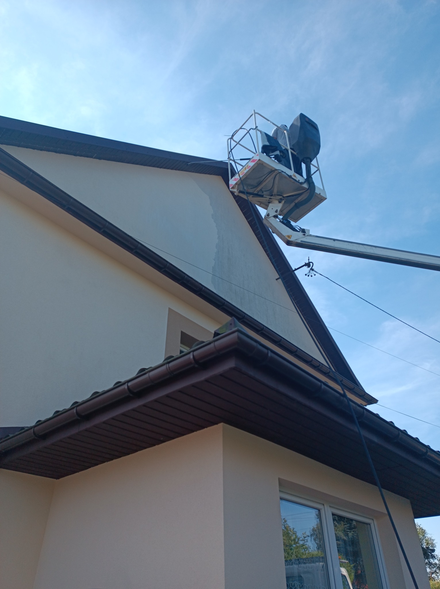 Pracownicy na podnośniku koszowym, w trakcie czyszczenia jasnej elewacji domu jednorodzinnego z brązowym dachem, widoczne smugi po myciu.