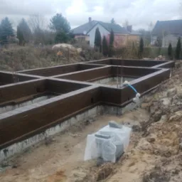 Zalane fundamenty z hydroizolacją w wykopie, zbrojenie pionowe. W tle zabudowa jednorodzinna i pochmurne niebo. Częściowo zakryte bloczki fundamentowe.