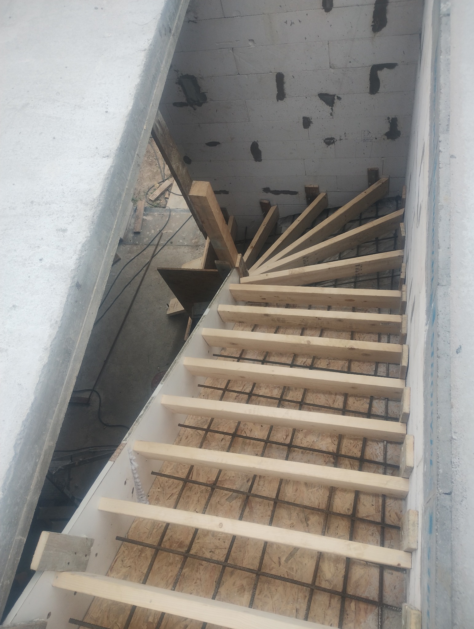 Wewnętrzna klatka schodowa w trakcie budowy. Widoczne zbrojenie, drewniane szalunki i stopnie. Ściany z betonu komórkowego. Ujęcie z góry, z widocznymi detalami konstrukcyjnymi.