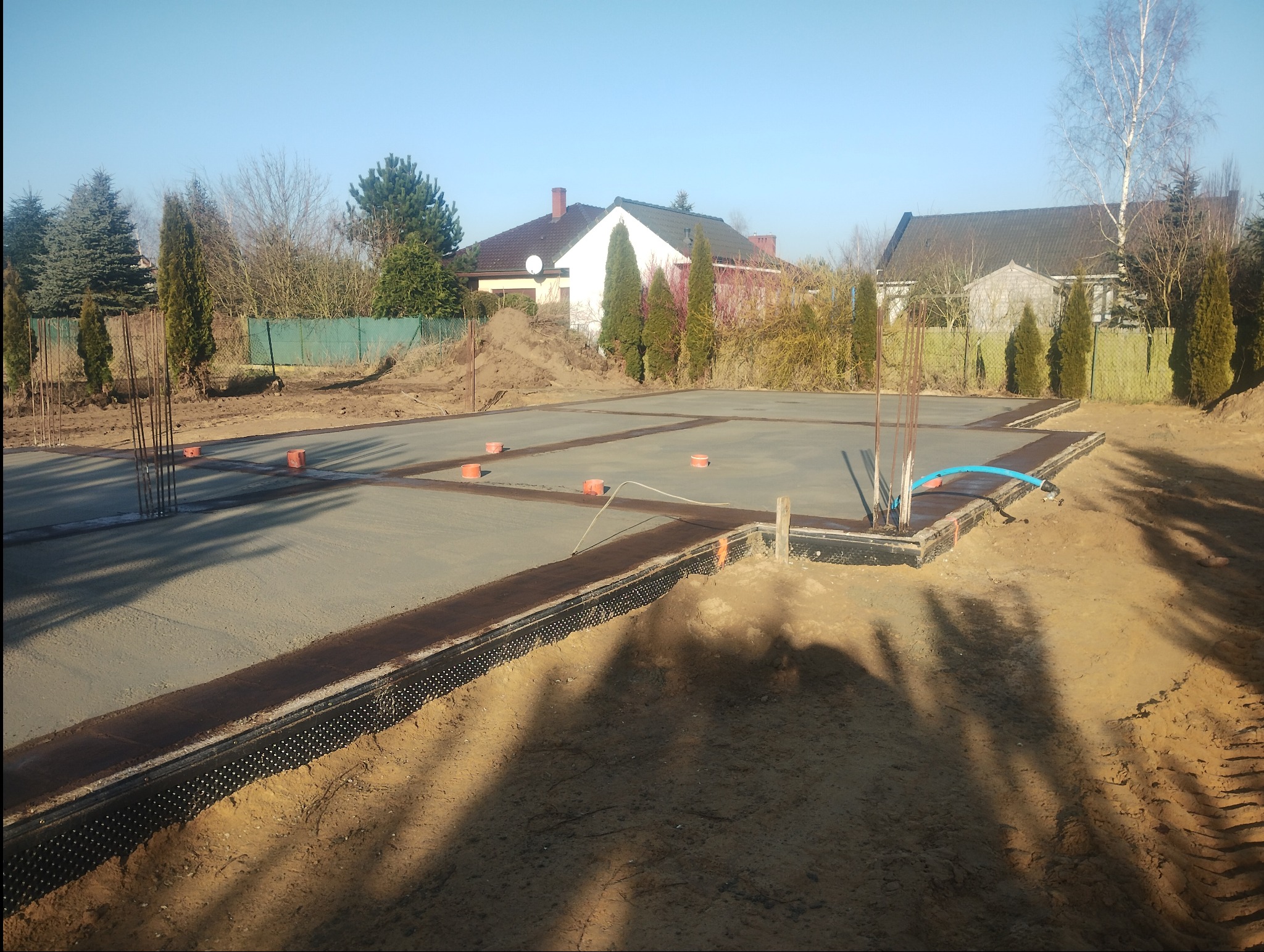 Wylana płyta fundamentowa z wystającymi prętami zbrojeniowymi na działce budowlanej. Widoczne deskowanie i izolacja pionowa. W tle zabudowa jednorodzinna.