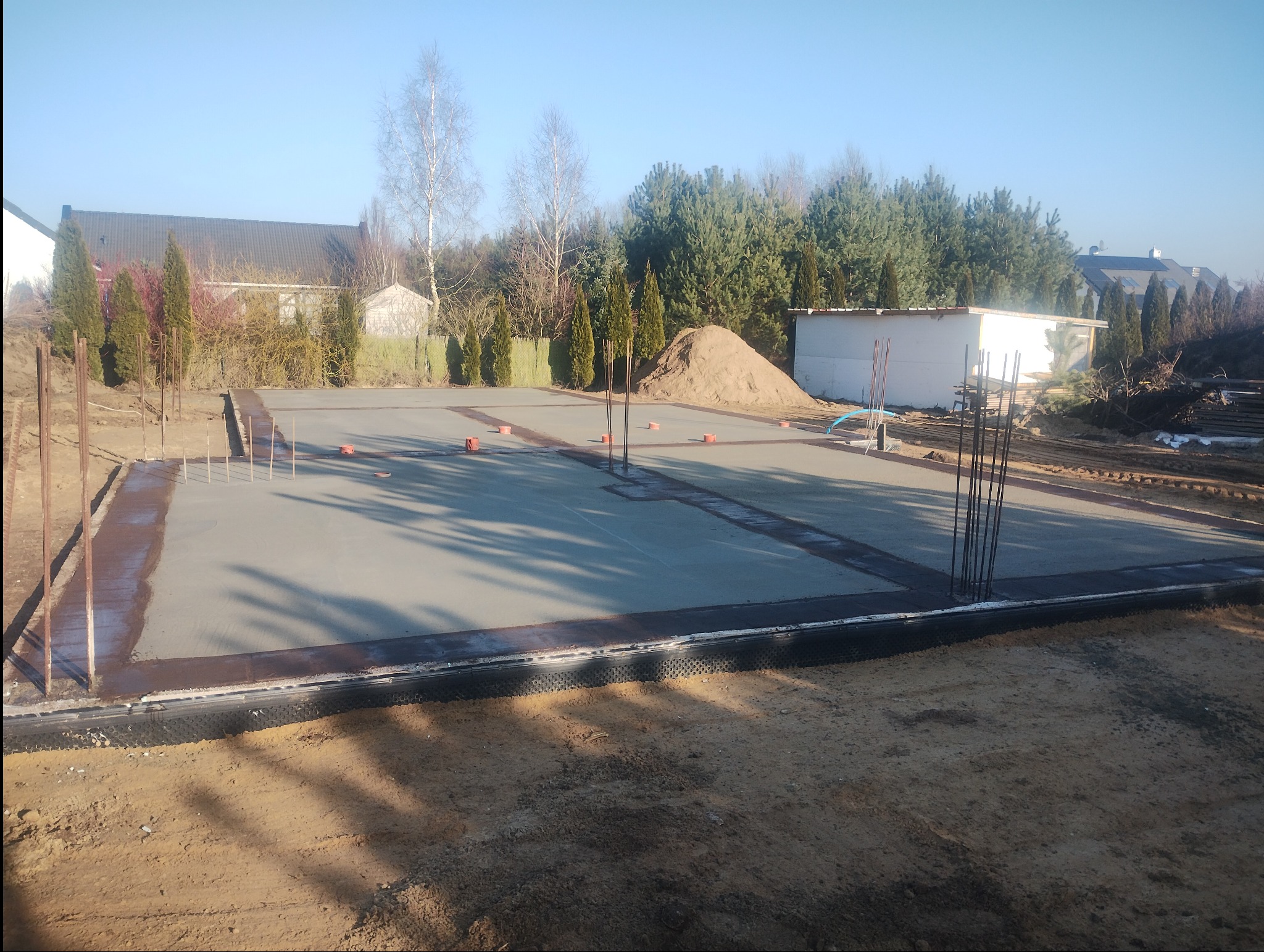 Wylany fundament pod budynek z wystającymi prętami zbrojeniowymi i widoczną izolacją przeciwwilgociową. W tle domy jednorodzinne i drzewa.