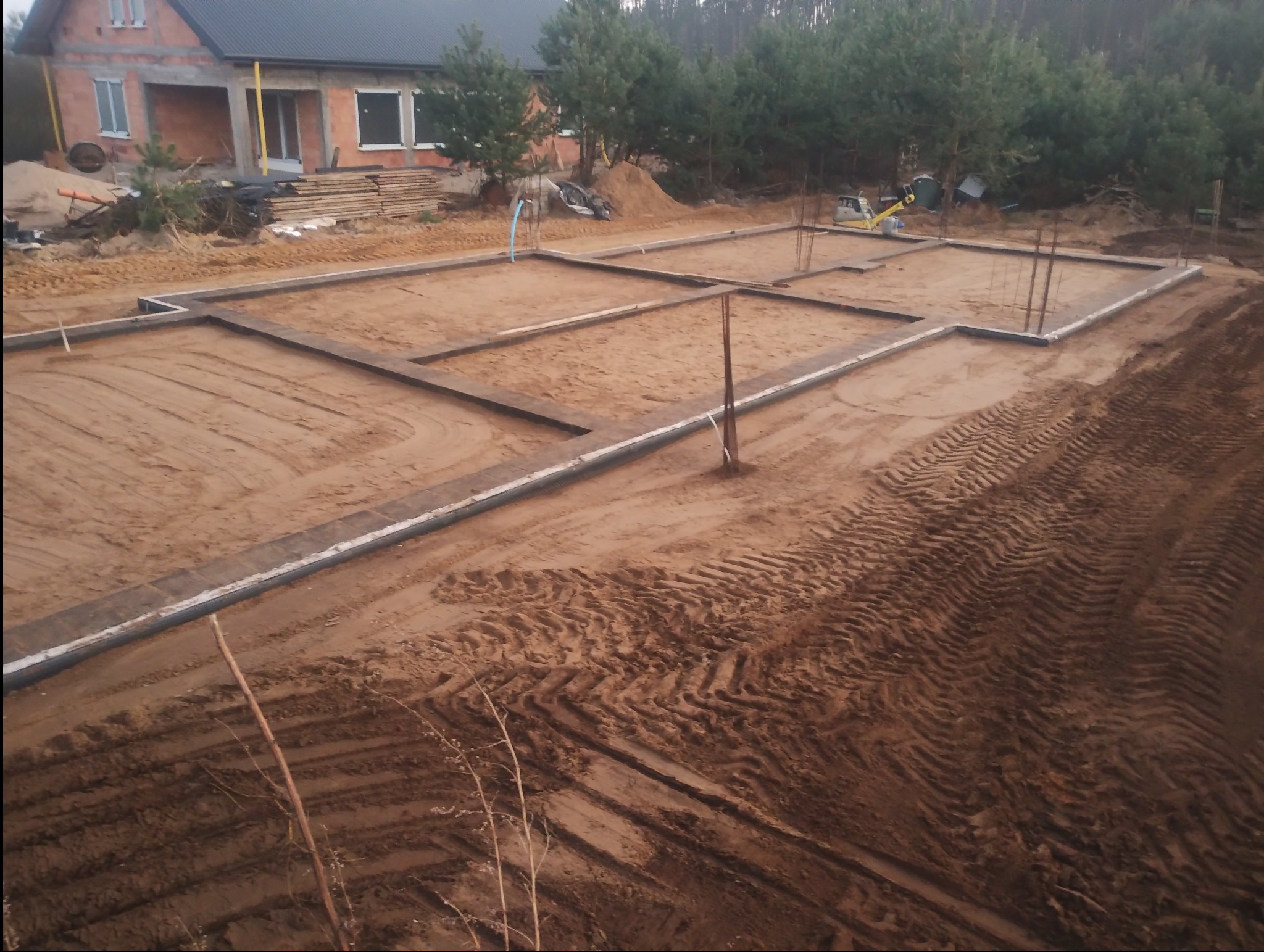 Zarys fundamentów domu jednorodzinnego z widocznymi szalunkami i zbrojeniami, w tle budynek w stanie surowym otwartym oraz drzewa.