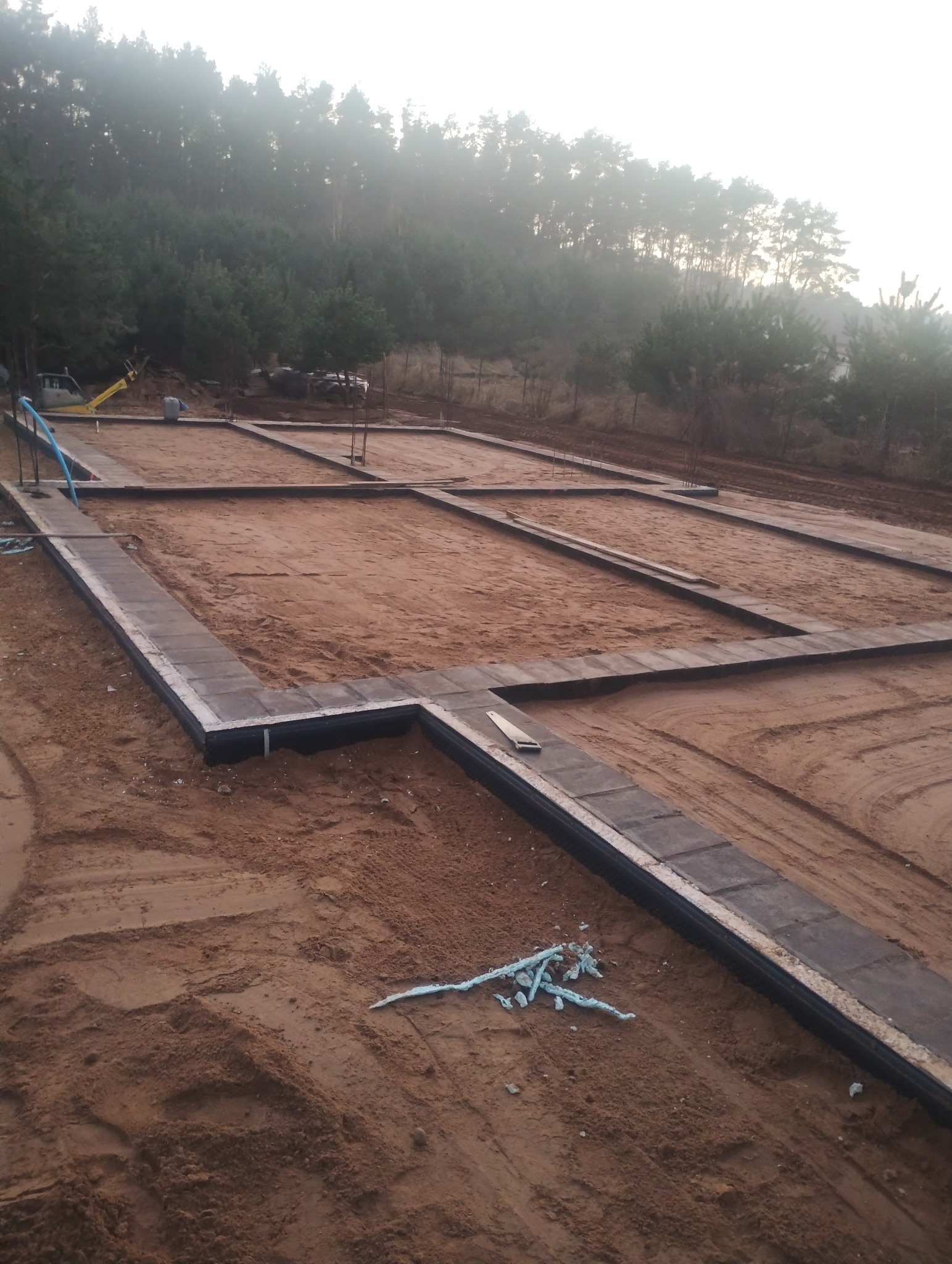 Zarys fundamentów domu jednorodzinnego na piaszczystym gruncie, widok z góry, w tle zalesione wzgórze. Etap przygotowania terenu pod budowę.