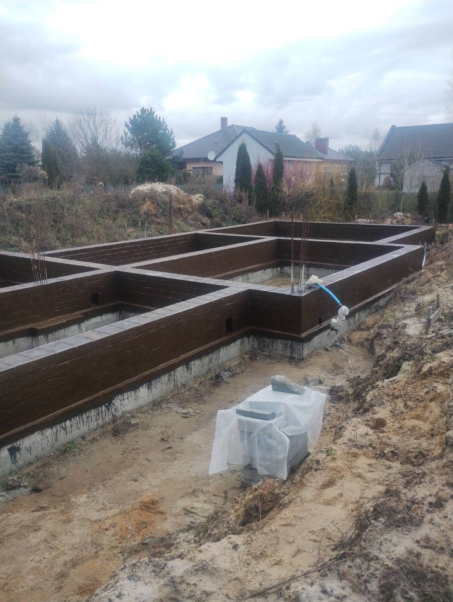 Zalane fundamenty z hydroizolacją w wykopie, zbrojenie pionowe. W tle zabudowa jednorodzinna i pochmurne niebo. Częściowo zakryte bloczki fundamentowe.