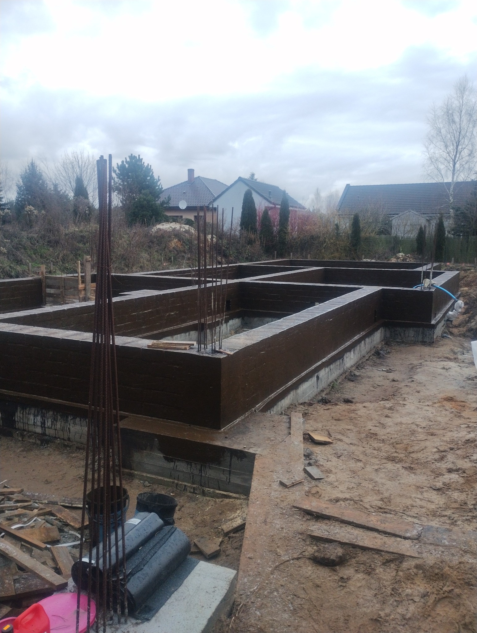 Zabezpieczone, brązową izolacją fundamenty pod budynek jednorodzinny. Widoczne wystające pręty zbrojeniowe i teren budowy w pochmurny dzień.