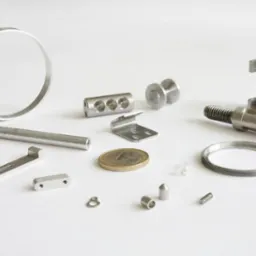 Precyzyjnie wykonane, metalowe elementy o różnych kształtach i rozmiarach, ułożone na białym tle, obejmujące pierścienie, pręty, blaszki i śruby, z monetą euro dla skali.