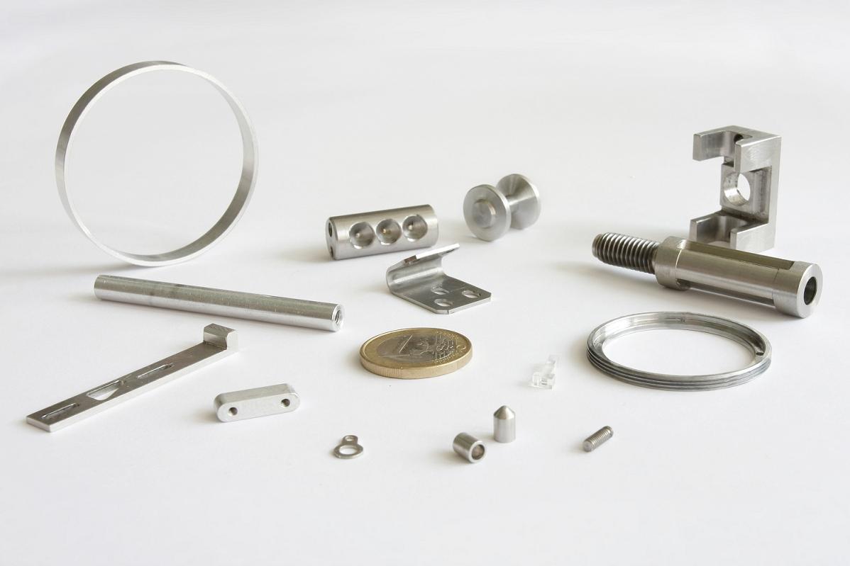 Precyzyjnie wykonane, metalowe elementy o różnych kształtach i rozmiarach, ułożone na białym tle, obejmujące pierścienie, pręty, blaszki i śruby, z monetą euro dla skali.