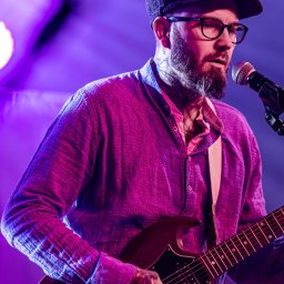 Seb Records - Wokalista z brodą i tatuażami na szyi, w kaszkiecie i okularach, grający na gitarze elektrycznej i śpiewający do mikrofonu, oświetlony fioletowym światłem scenicznym.