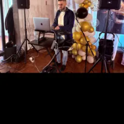 DJ z brodą i okularach gra na keyboardzie Recordbox na tle dekoracji z białych i złotych balonów oraz dwóch głośników JBL podczas imprezy.