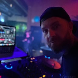 Seb Records - DJ w czapce za konsoletą mikserską w klubie, oświetlony neonami, z zamyślonym wyrazem twarzy, w tle rozmazane sylwetki bawiących się osób.