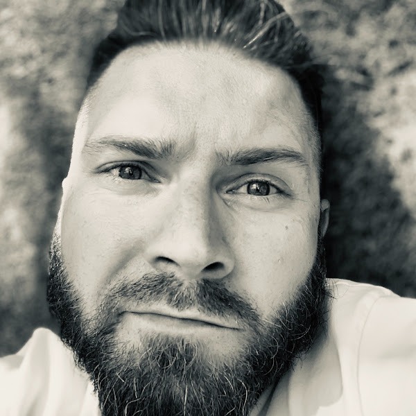 Czarno-biały portret brodatego mężczyzny z bliska, z delikatnym uśmiechem i intensywnym spojrzeniem w obiektyw.