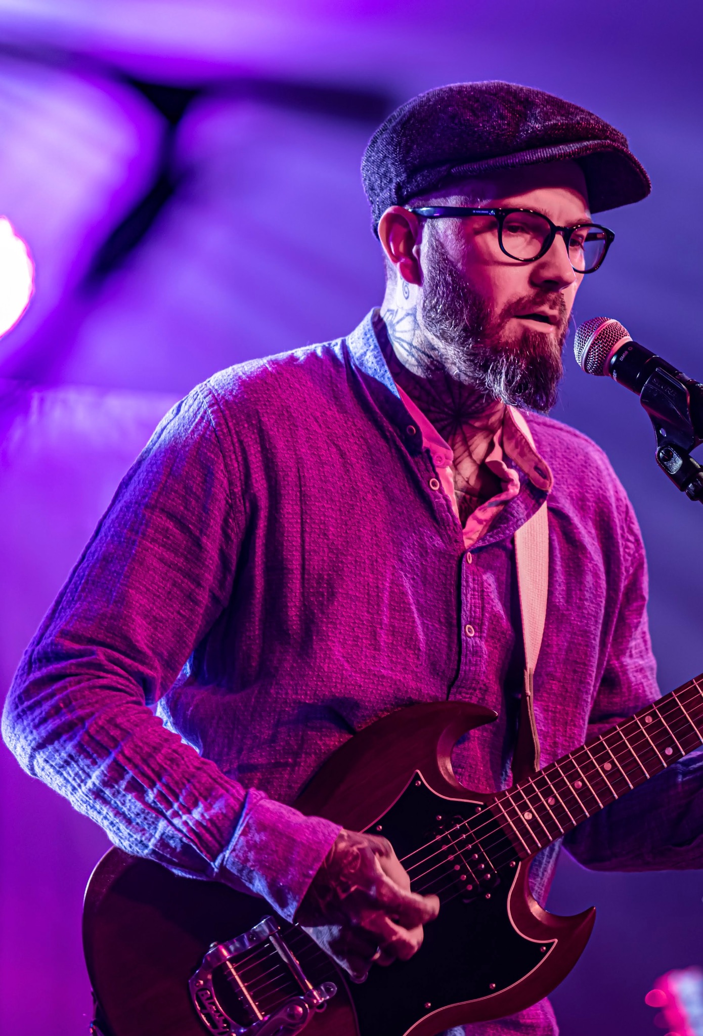 Wokalista z brodą i tatuażami na szyi, w kaszkiecie i okularach, grający na gitarze elektrycznej i śpiewający do mikrofonu, oświetlony fioletowym światłem scenicznym.