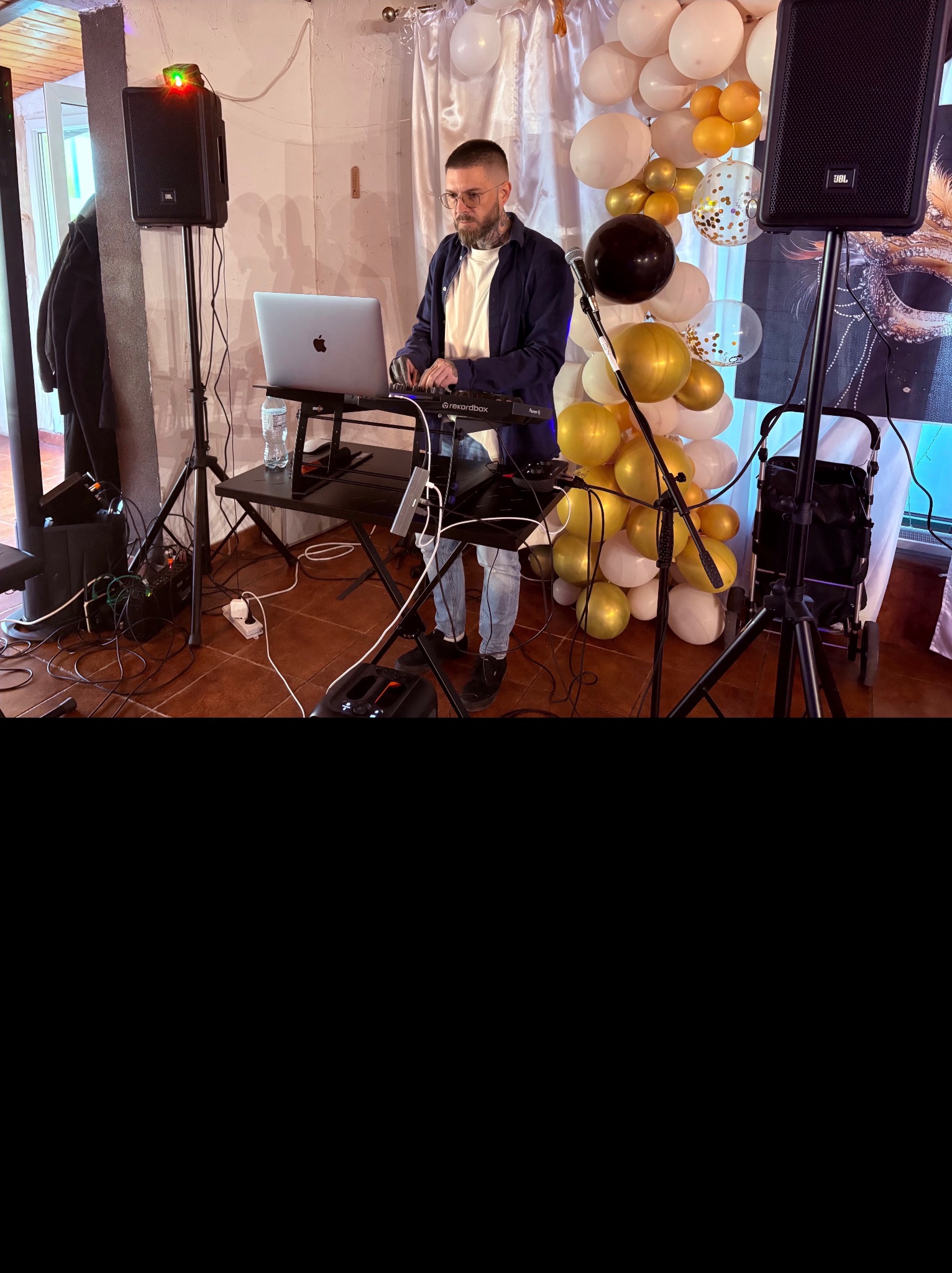DJ z brodą i okularach gra na keyboardzie Recordbox na tle dekoracji z białych i złotych balonów oraz dwóch głośników JBL podczas imprezy.