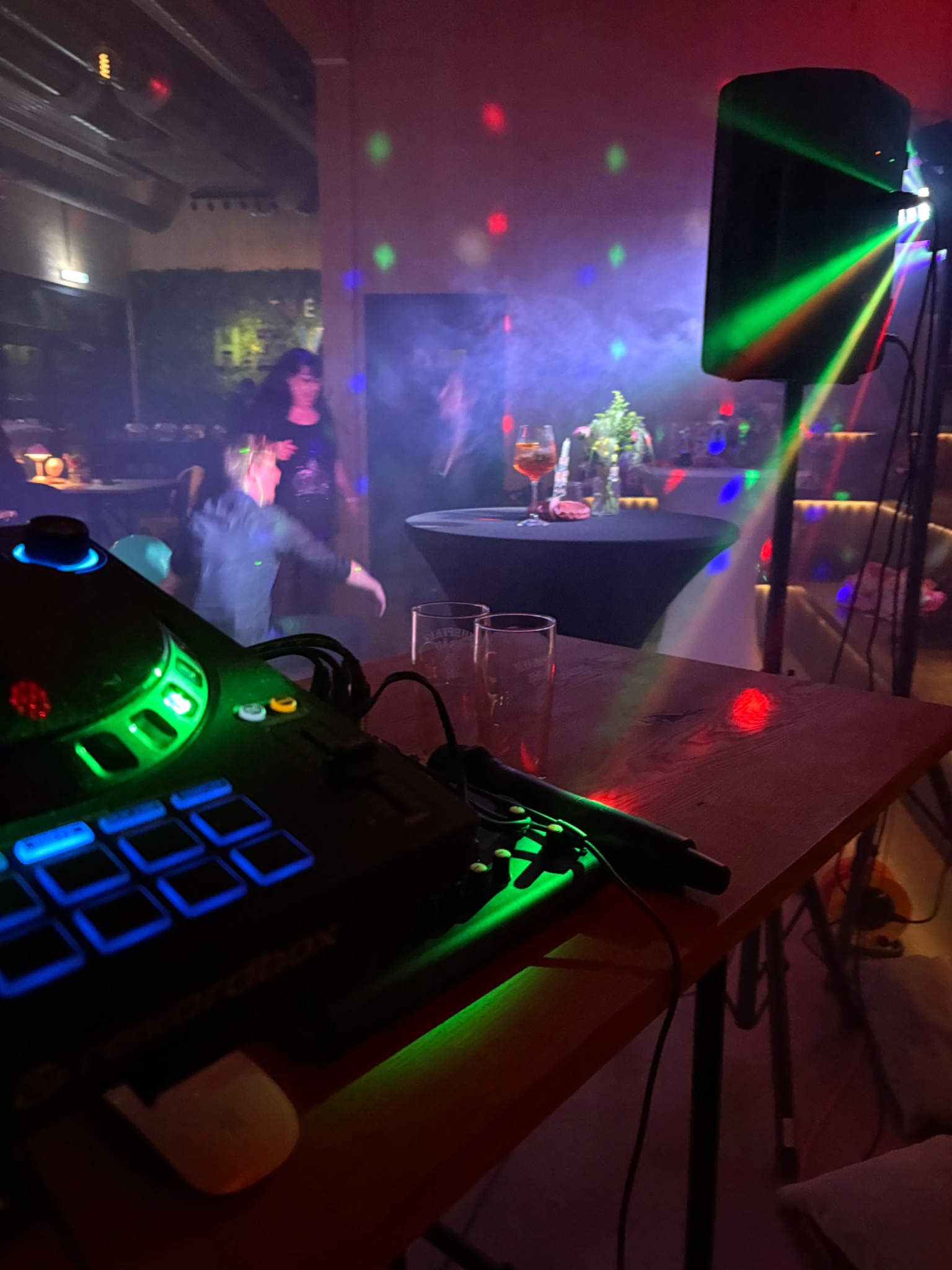 Oświetlona scena z DJ-em, sprzętem audio, dekoracjami i tańczącymi osobami w tle, tworząca imprezową atmosferę. Laserowe światła i efekty dymne.