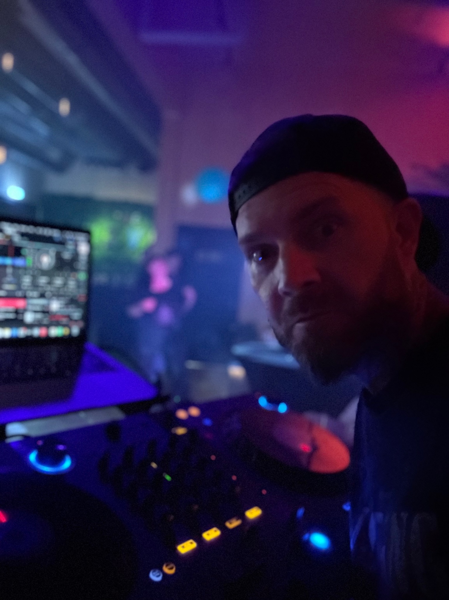 DJ w czapce za konsoletą mikserską w klubie, oświetlony neonami, z zamyślonym wyrazem twarzy, w tle rozmazane sylwetki bawiących się osób.