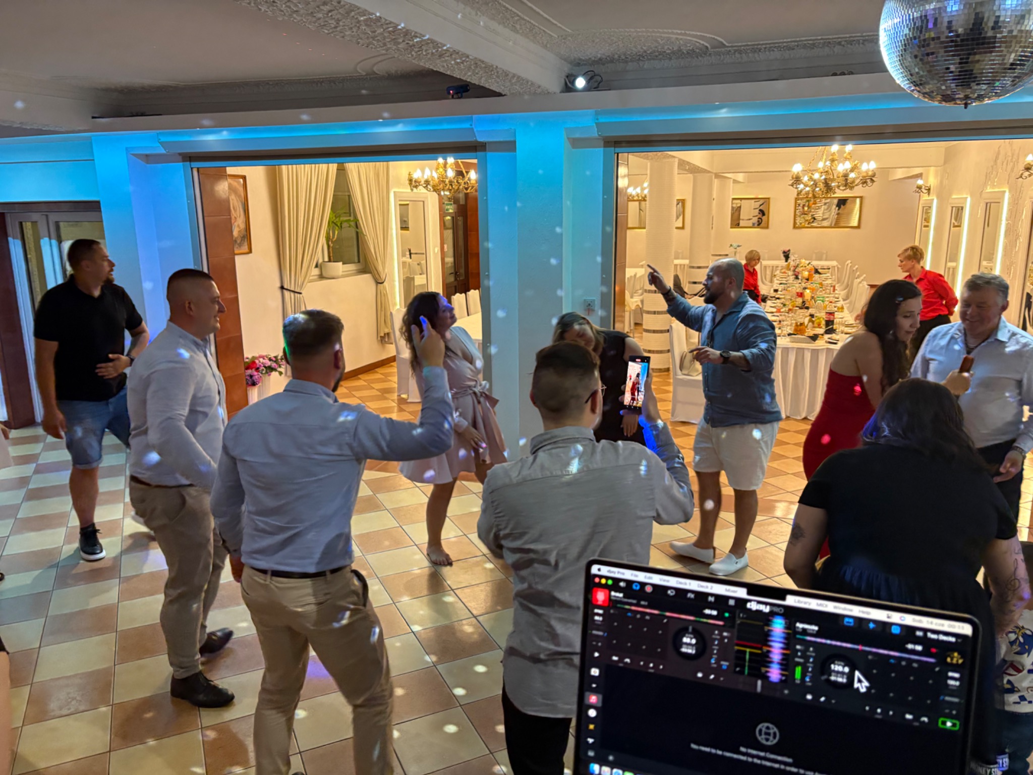 Roztańczona grupa gości weselnych w sali balowej z DJ-em. Laptop z programem DJ-skim na pierwszym planie, parkiet w tle, światła dyskotekowe.