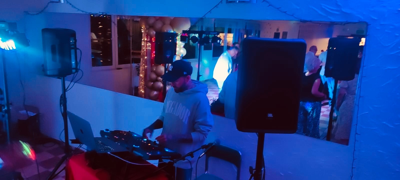 DJ w trakcie występu na imprezie, widoczne konsoleta, głośniki i bawiący się ludzie w tle, odbici w lustrze. Scena oświetlona niebieskim światłem.