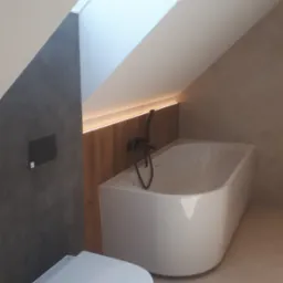 Nowoczesna łazienka na poddaszu ze skośnym sufitem, biała wanna z czarną armaturą, podwieszana toaleta, szara ściana i oświetlenie LED za drewnianą boazerią.